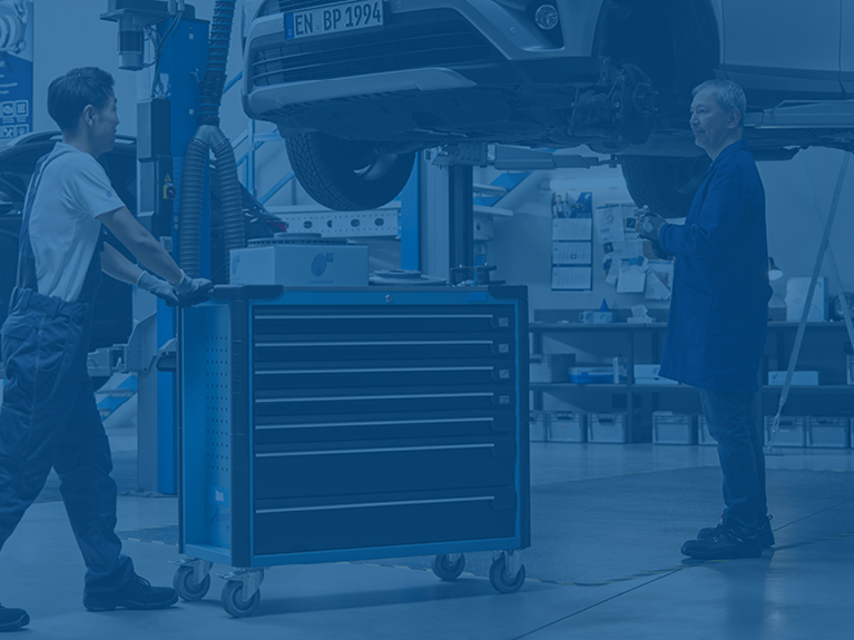 Автомеханік підкочує візок з інструментами та запчастинами до піднятого на підйомнику азіатського автомобіля, де другий механік очікує на запасну частину