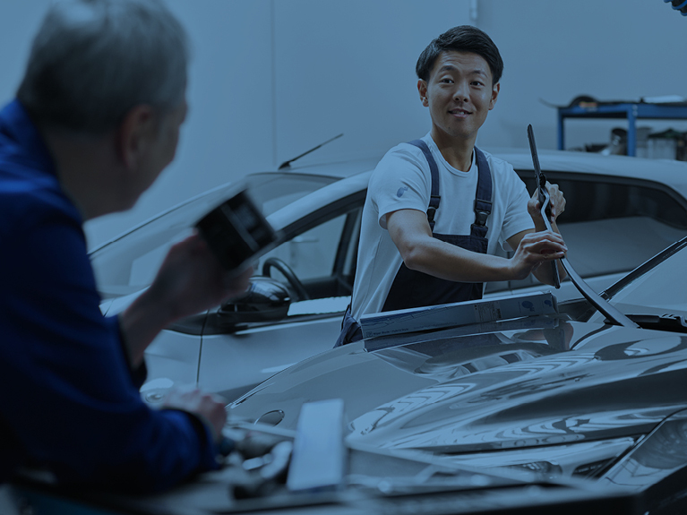 Два механіка нахилились над закритим капотом, один з них встановлює щітки склоочисників